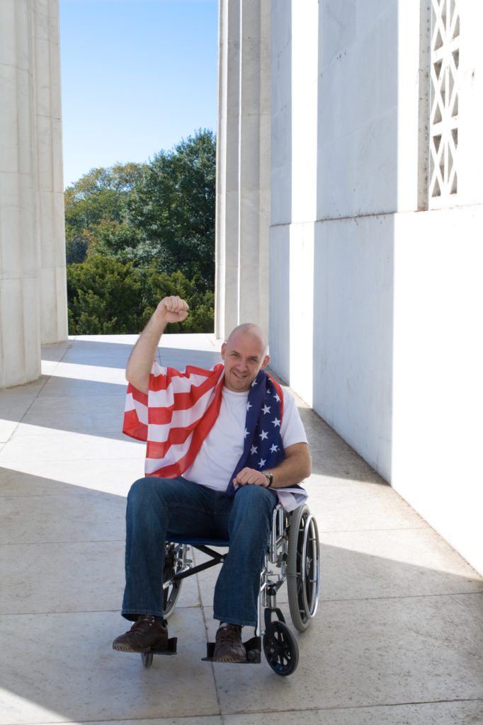 Patriotic white man sitting in a wheelchair with raised fist and American flag draped across his shoulders. Shot at the Lincoln Memorial, in Washington, DC, USA.
