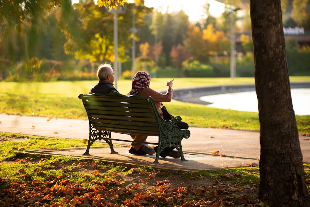 Elderly couple sitting on park bench, enjoying autumn view with fallen leaves.